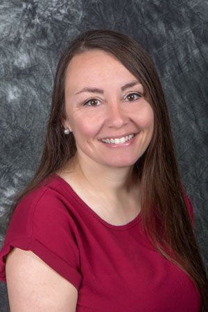 Woman with long brown hair smiling, wearing a red top, against a mottled gray backdrop.