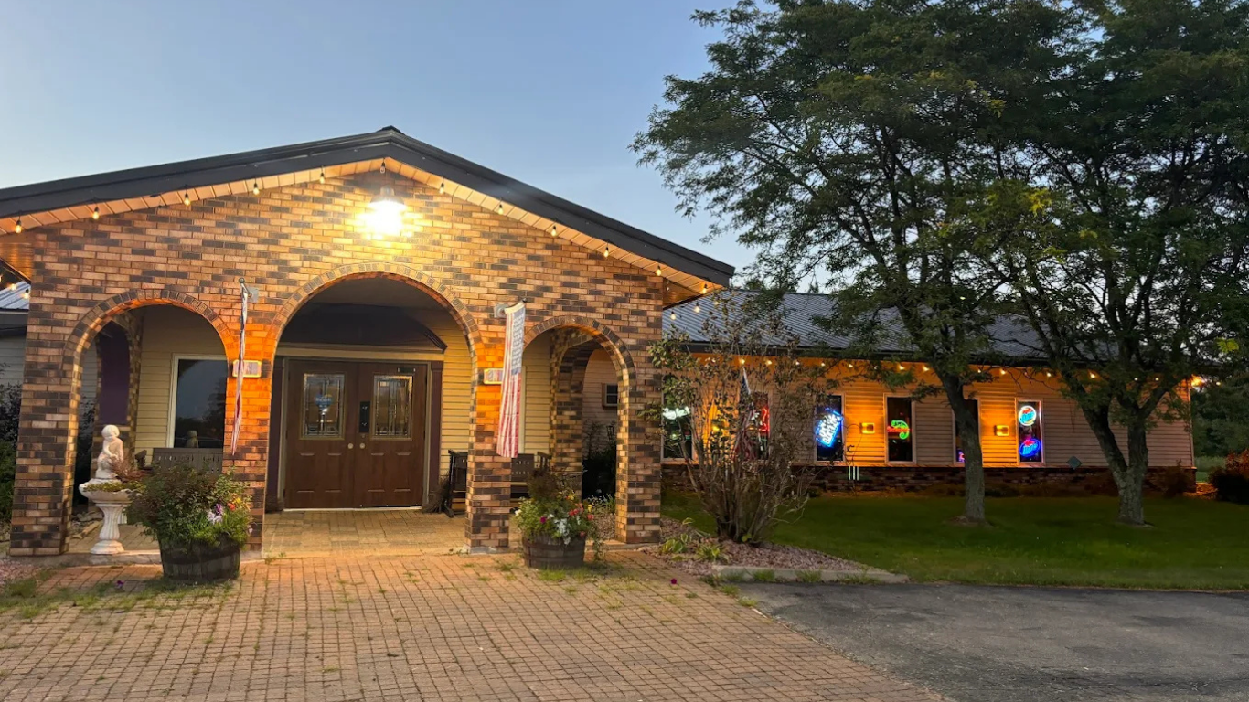 Stone-faced building with arched entrance and side windows. Lit by outdoor lights. A tree and grassy area are beside the structure.
