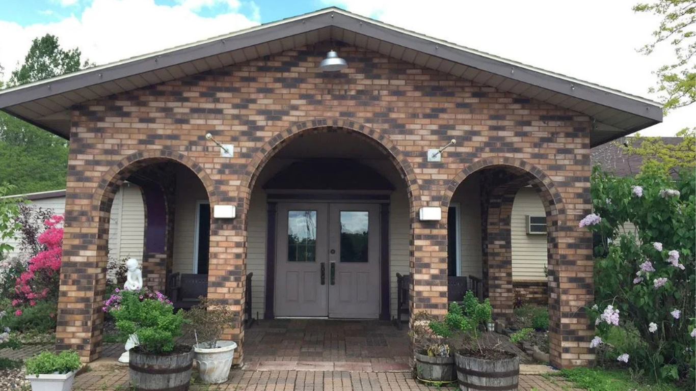Brick building with arched entrance and double doors, greenery and potted plants in front.