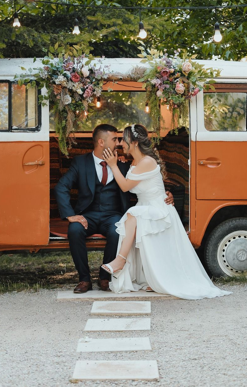 Couple in wedding attire embrace in an orange van, decorated with flowers and lights.