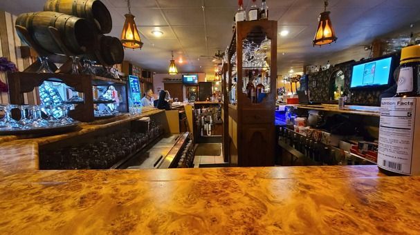 Bar interior with wooden bar top, ornate carvings, barrels, and liquor bottles.