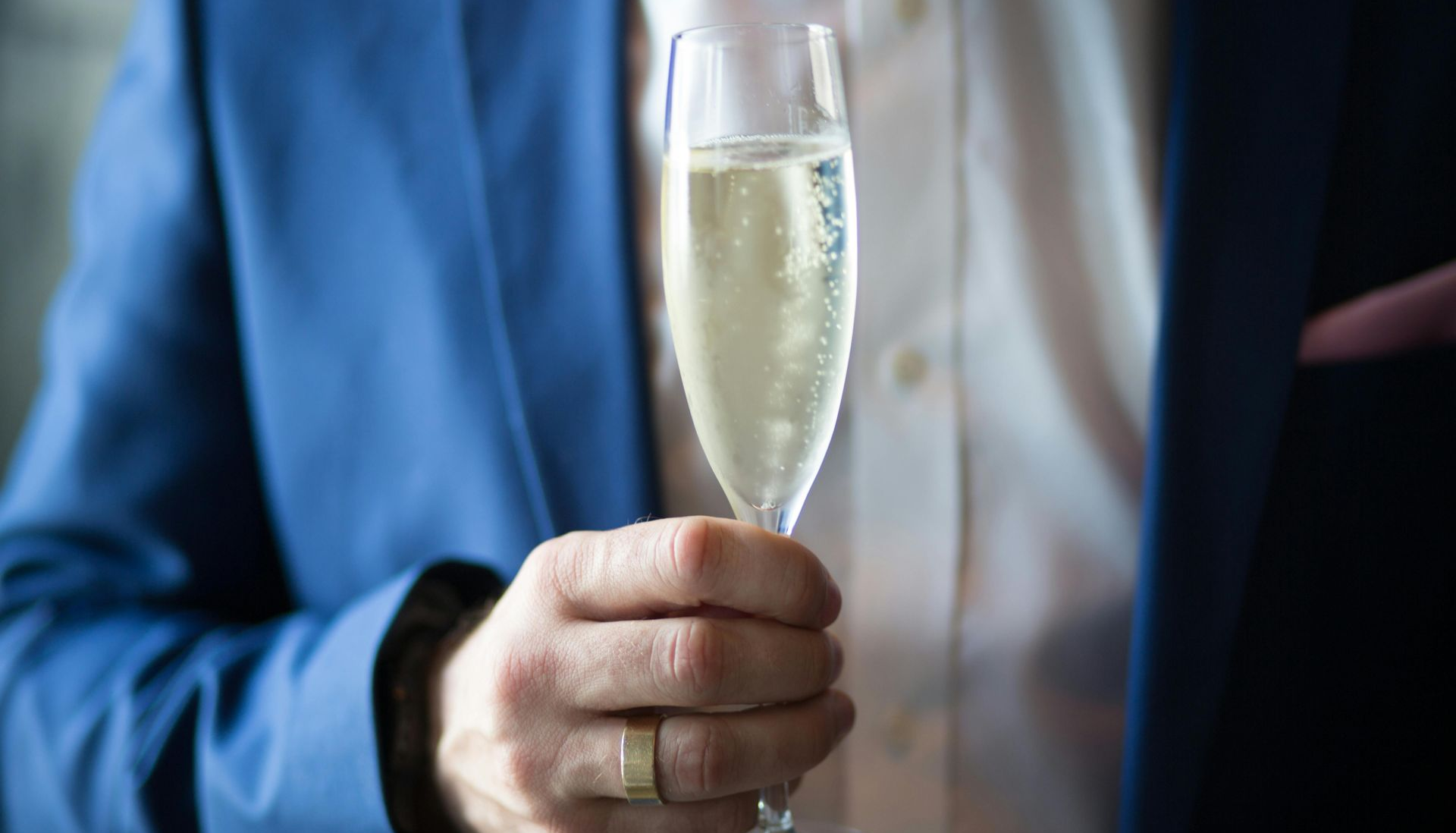 Person in blue blazer holding champagne flute, gold ring visible.
