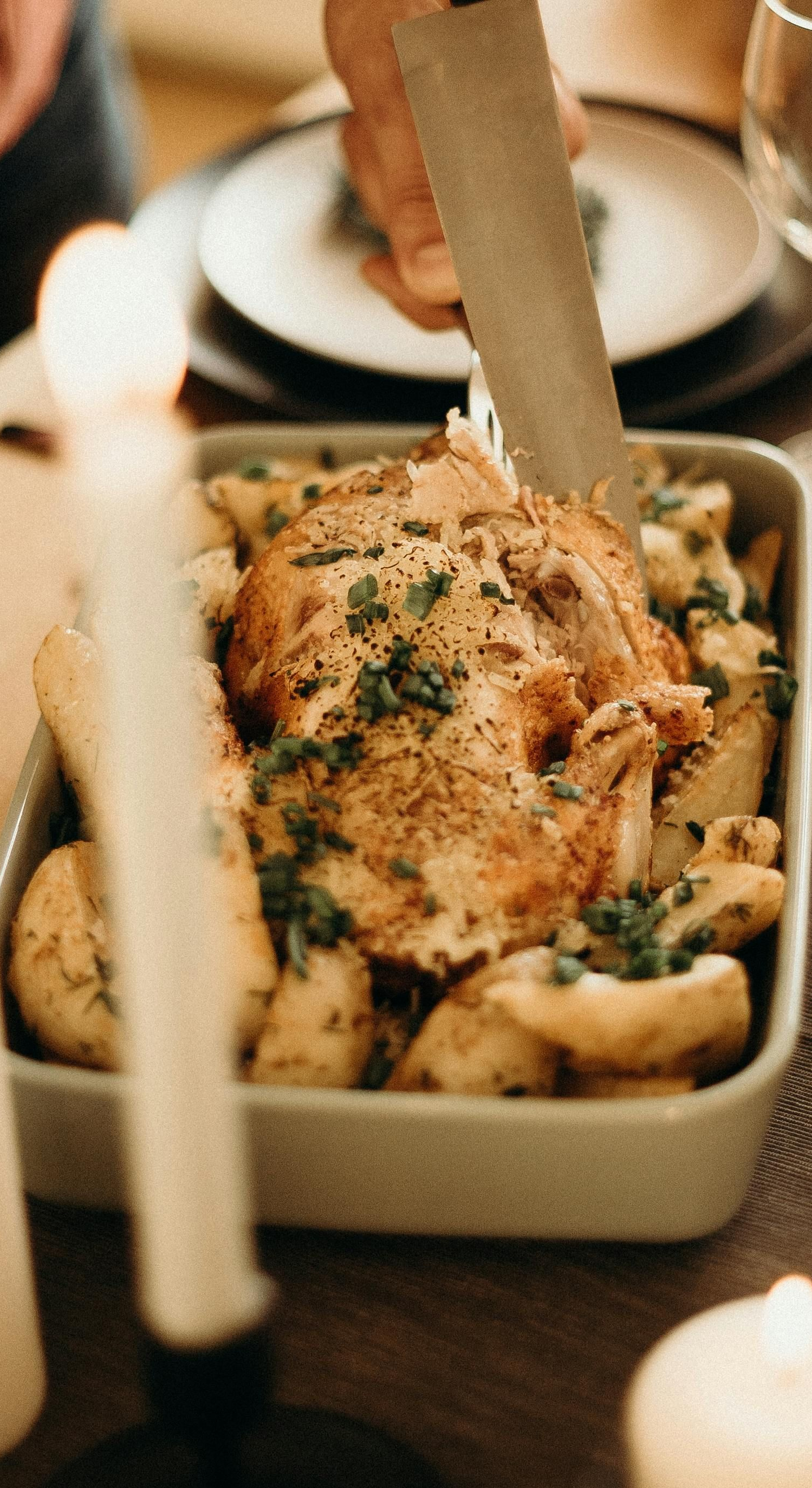 Roast chicken with potatoes in a baking dish, being carved at a dinner table.