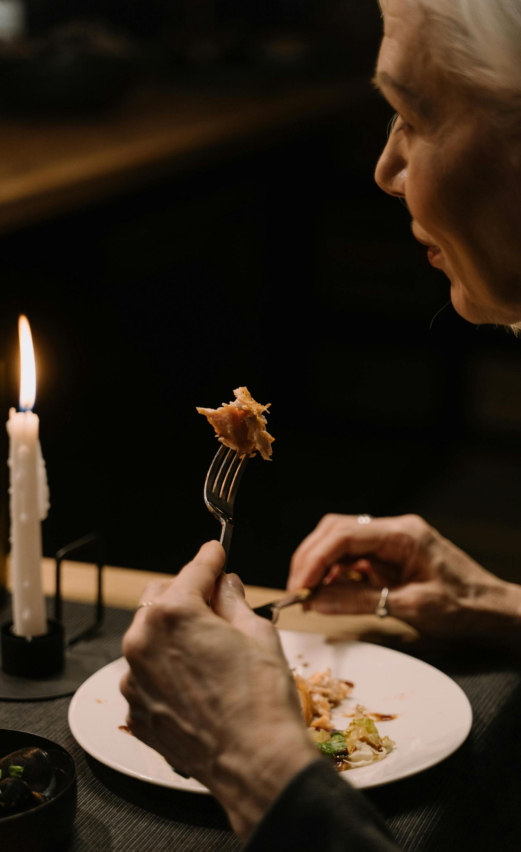 Person eating a meal by candlelight, holding a fork with food over a white plate.