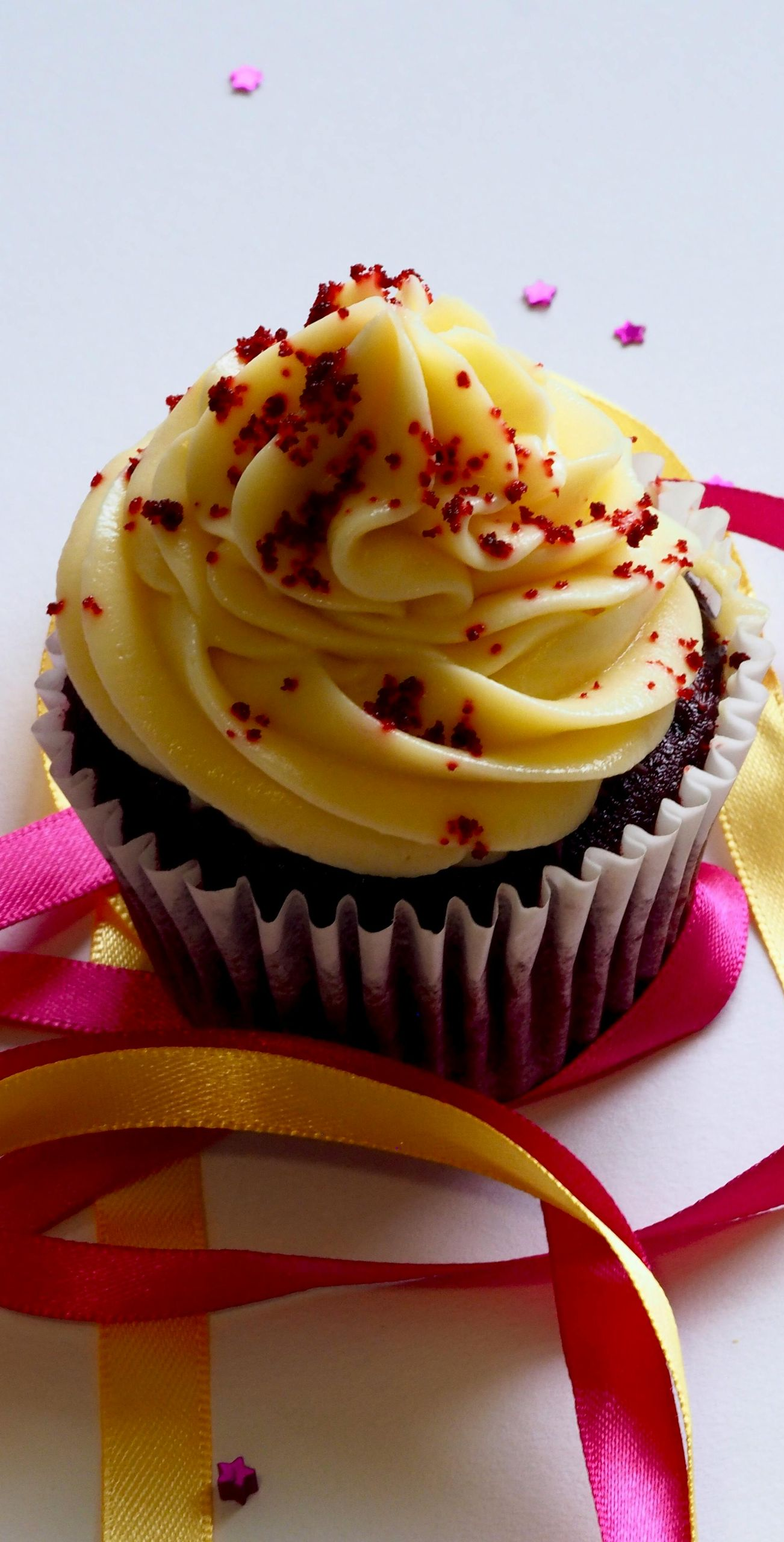 Red velvet cupcake with yellow frosting, sprinkled with red crumbs, sits on ribbons.