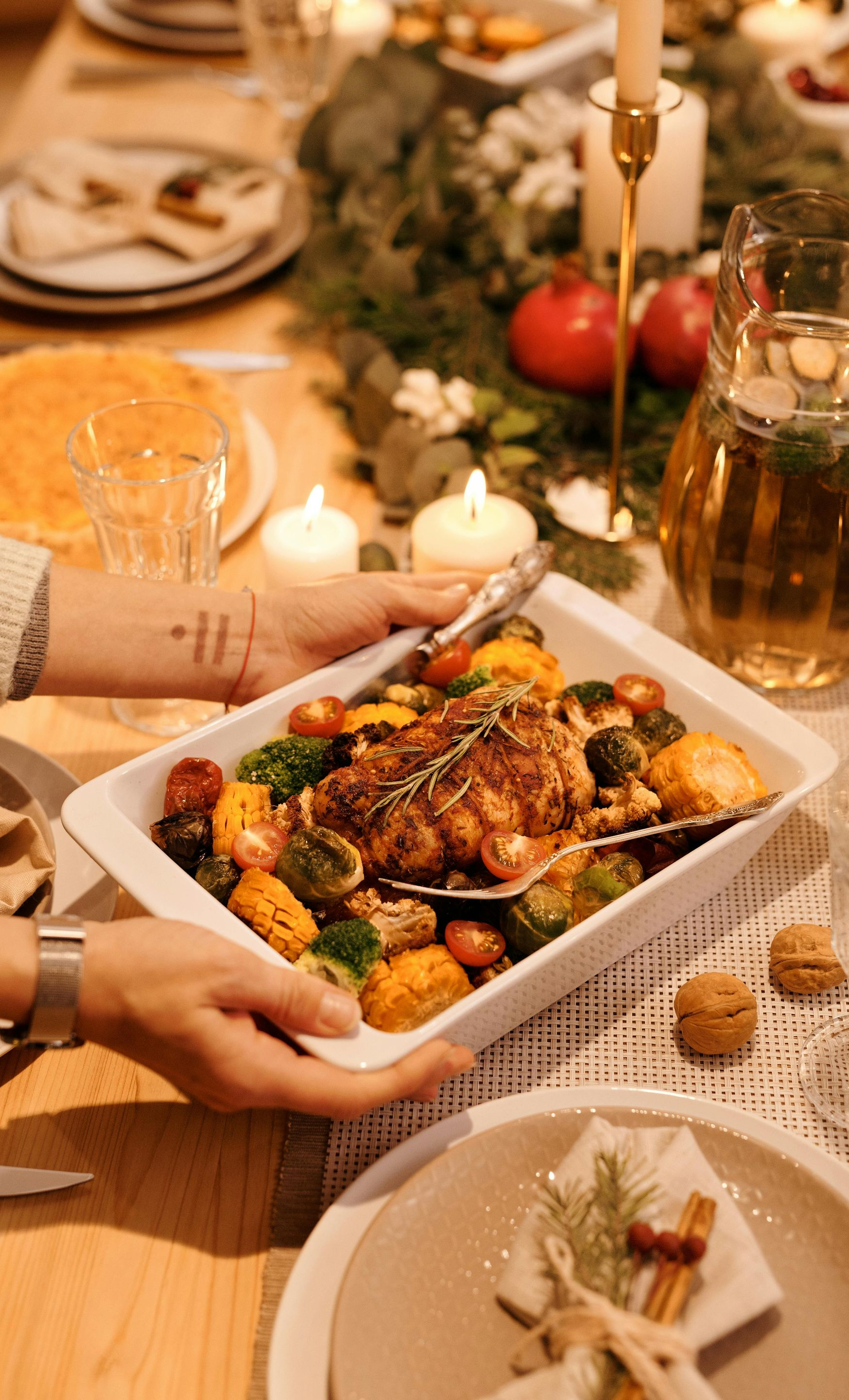 Person holding a baked dish of roasted vegetables and meat at a festive dinner table setting.
