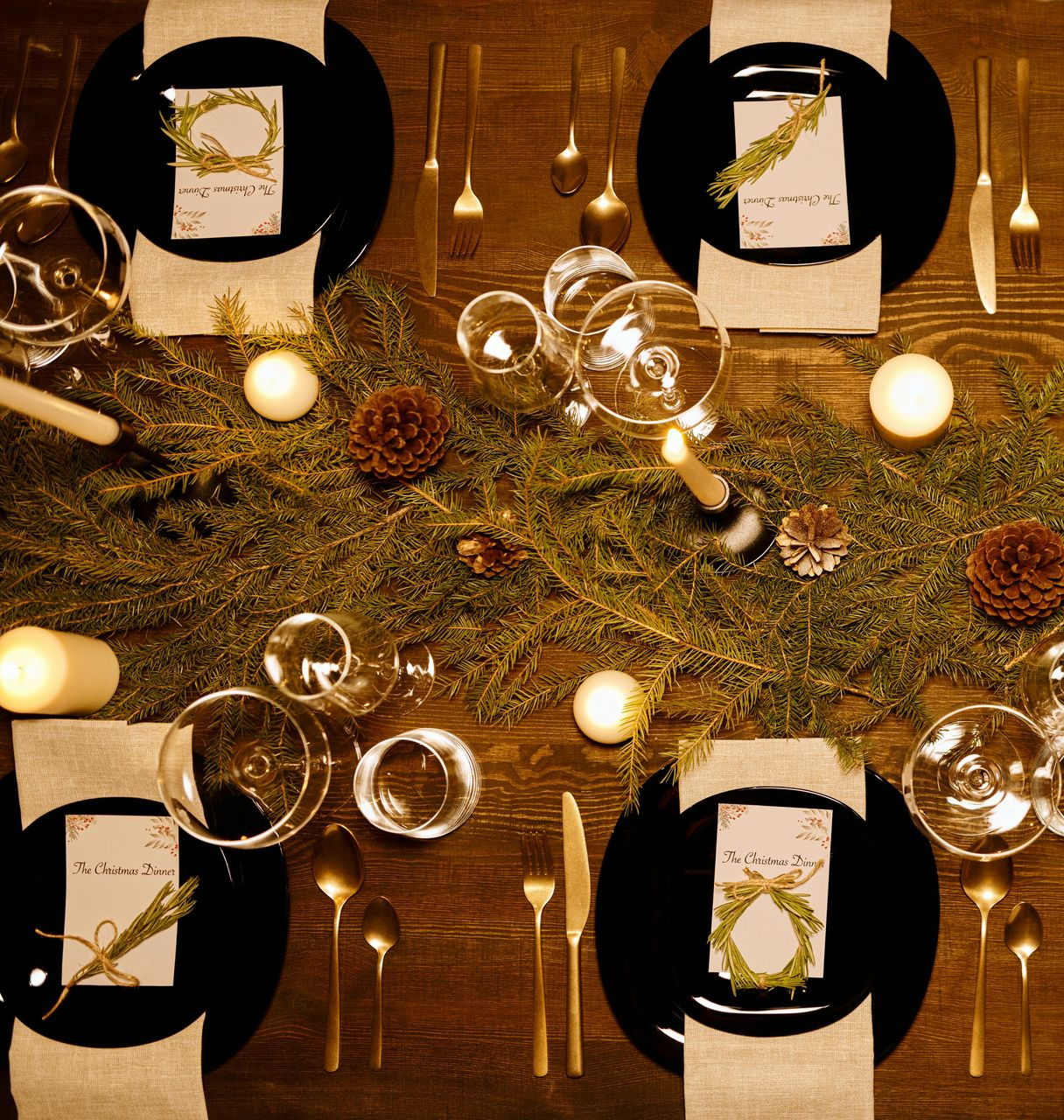 Elegant dining table set with black plates, gold cutlery, candles, and greenery.