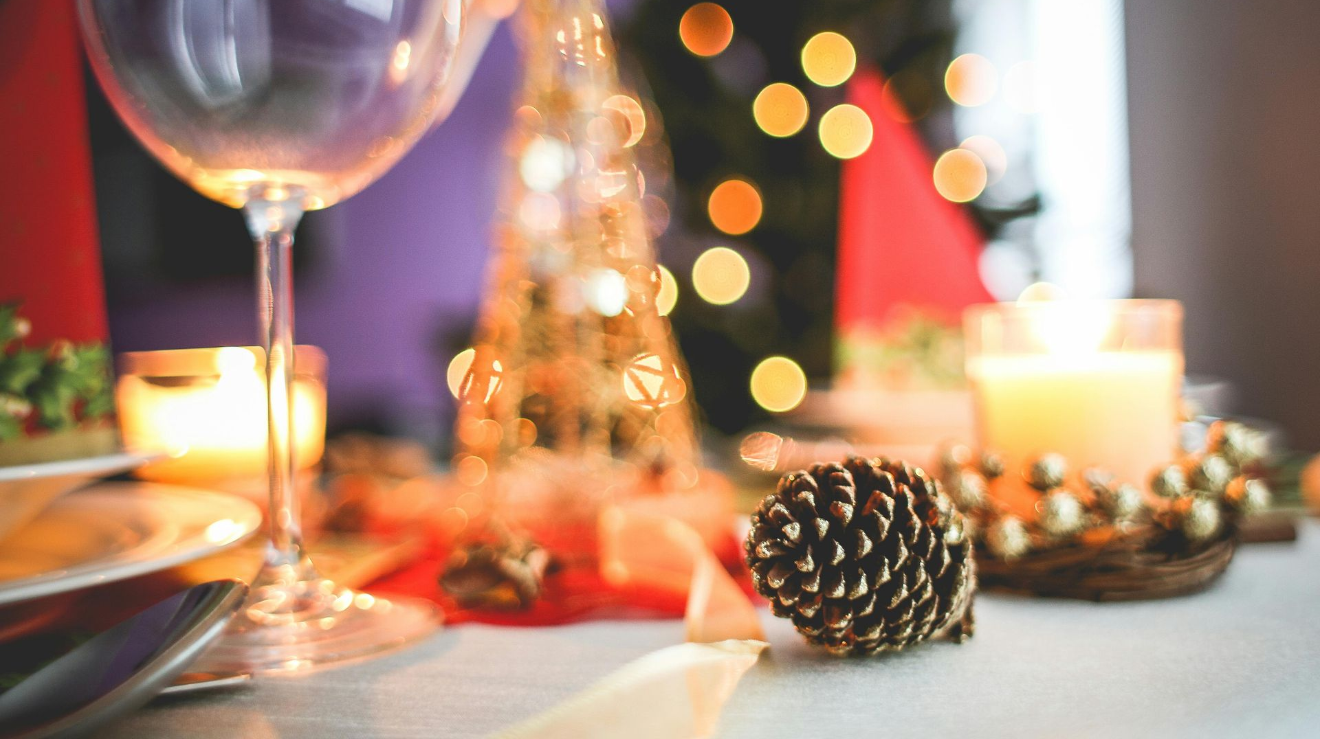 Festive table setting with wine glass, candle, pinecone, and blurred Christmas lights.