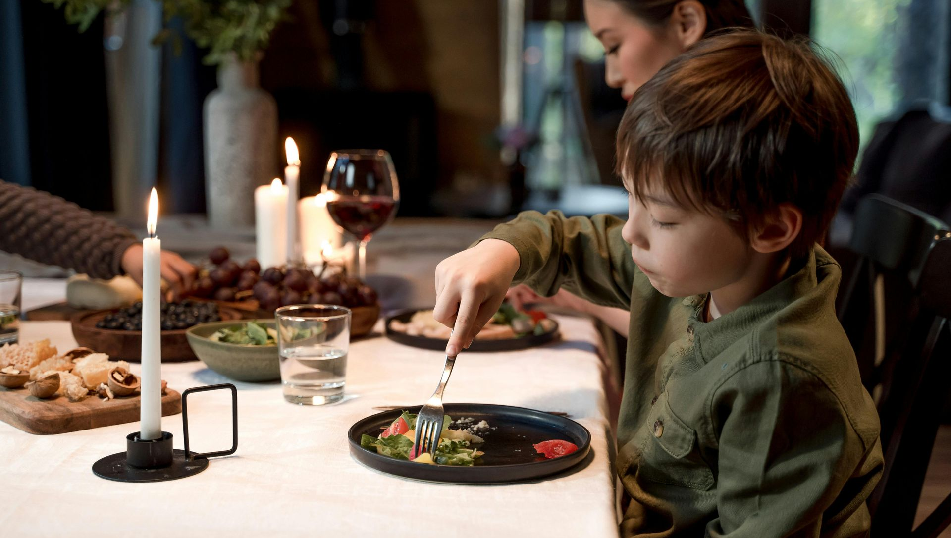 Child at dinner table, cutting food with fork and knife, lit by candlelight.