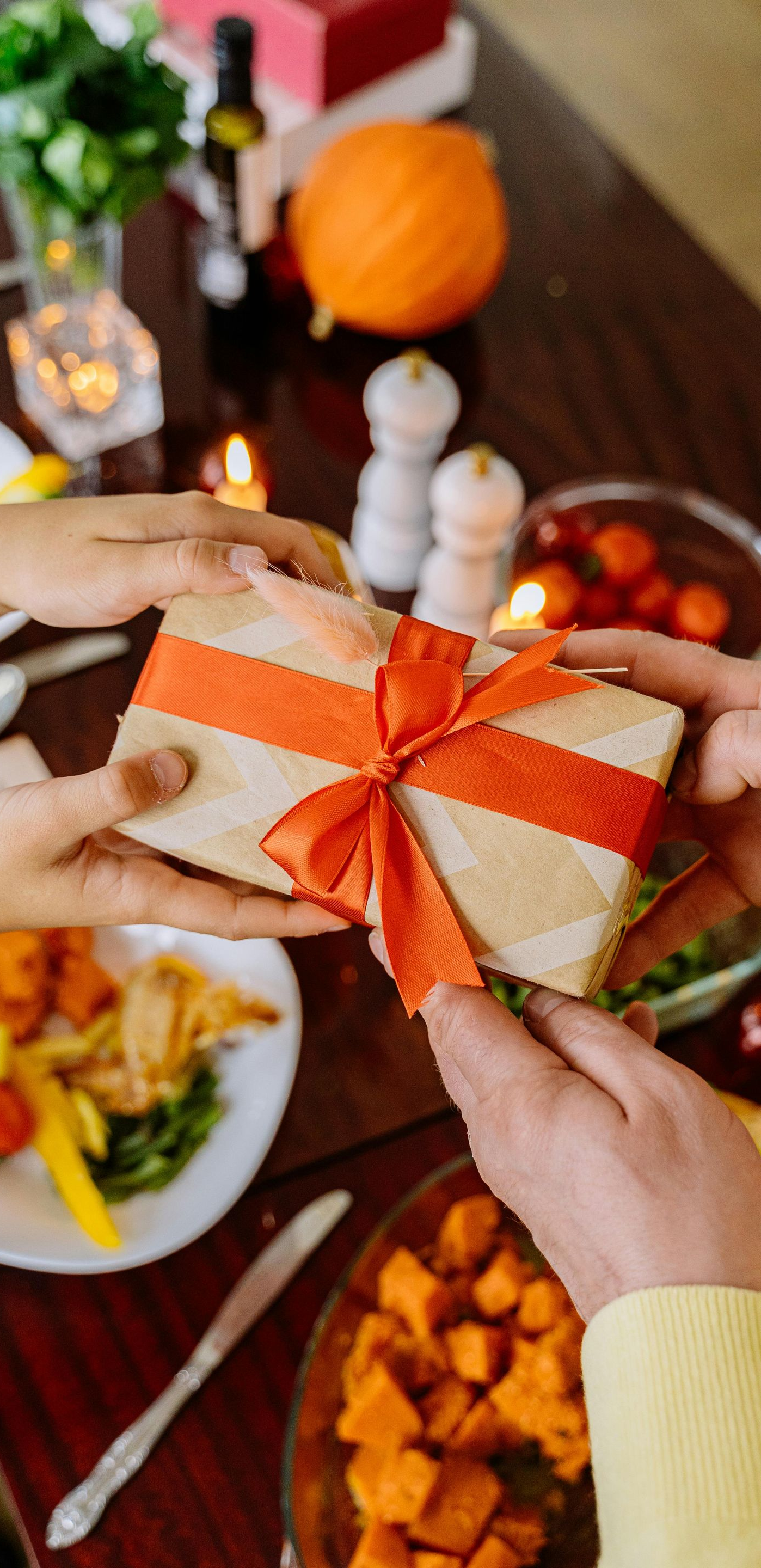 Hands passing a wrapped gift with an orange ribbon over a holiday meal.