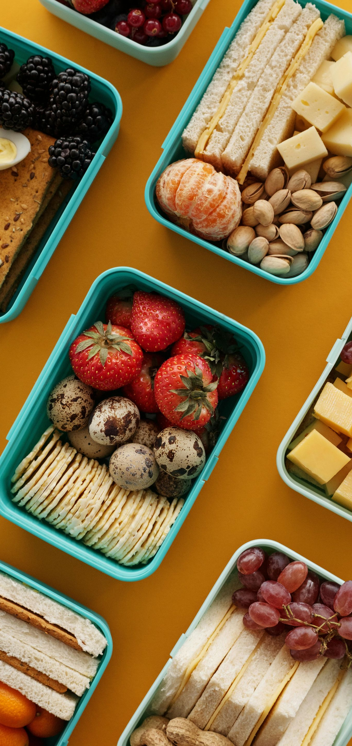Several turquoise lunchboxes filled with different snacks, arranged on a yellow surface.