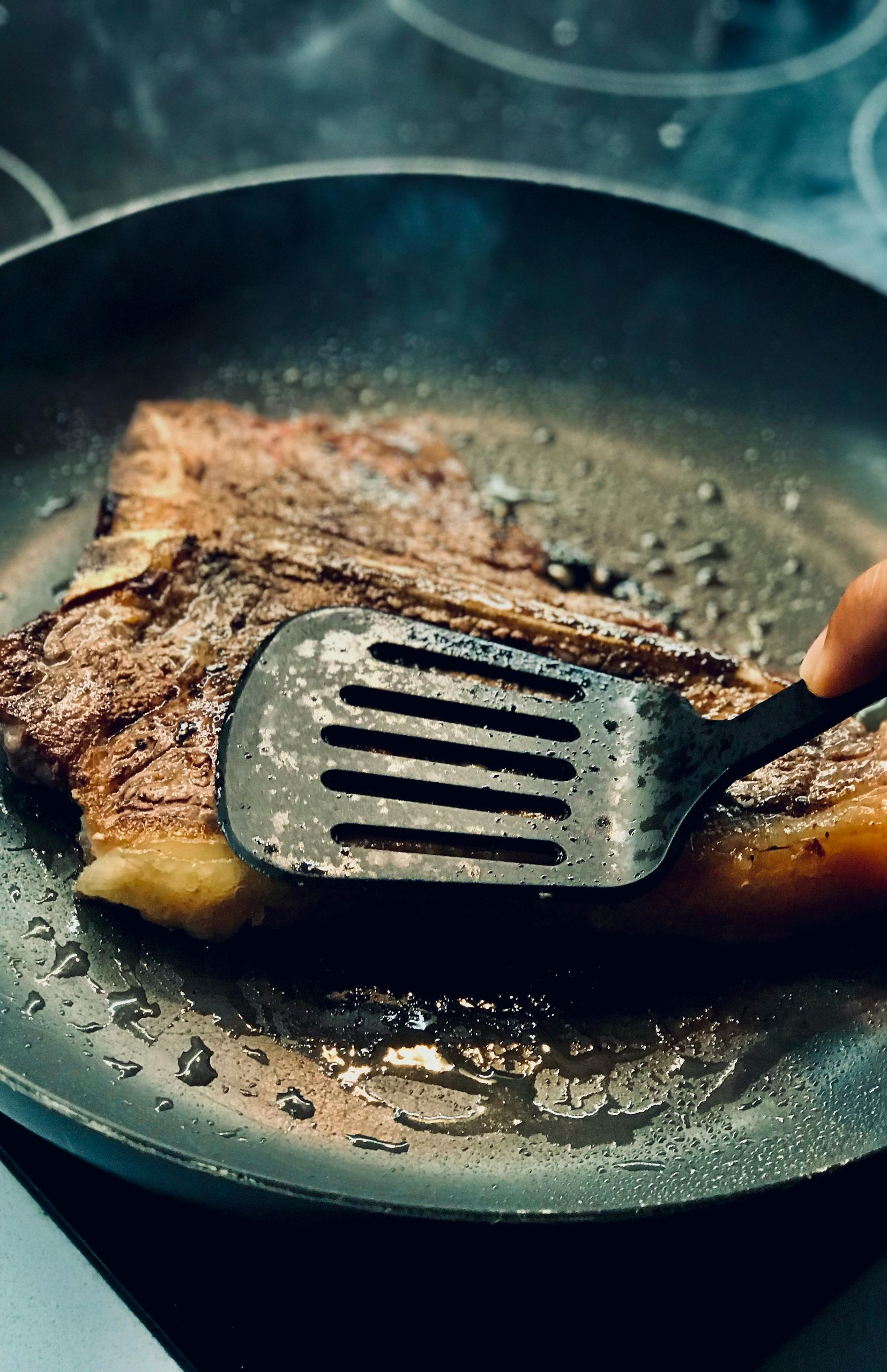 A steak being cooked in a pan; a hand uses a spatula to flip it.