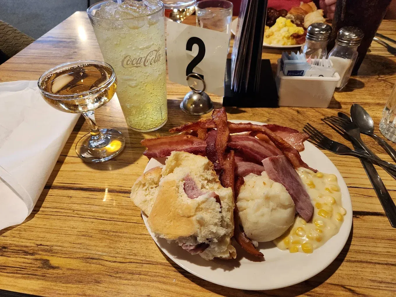 Plate of food with bacon, bread, mashed potatoes, and corn at a table setting. Drinks and table number visible.