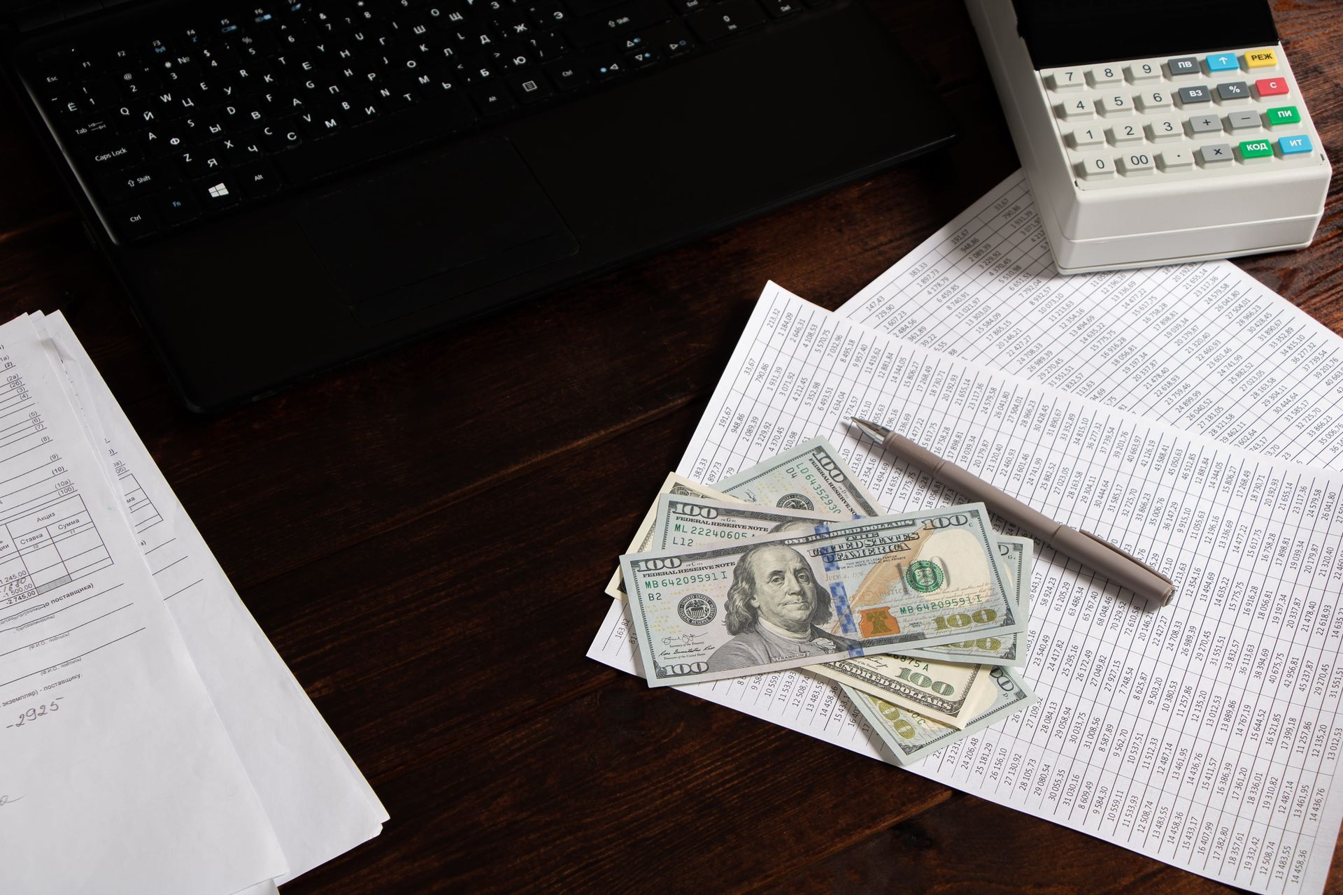 A laptop, a numeric keypad, a pen, and US dollar bills resting on printed documents on a dark wooden desk.