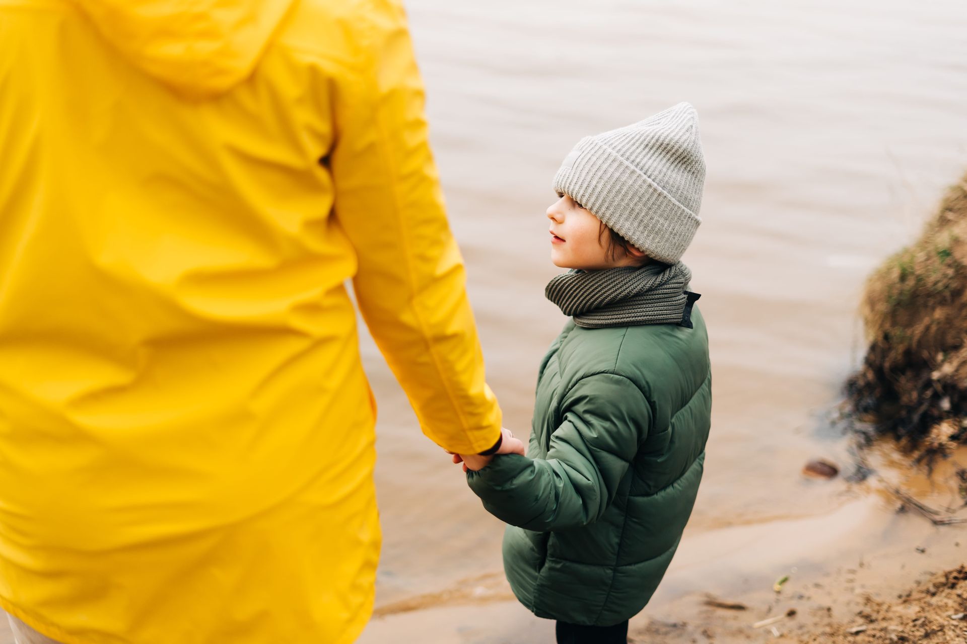 A person in a yellow raincoat holds hands with a child in a green puffer jacket and gray beanie near a sandy shore.