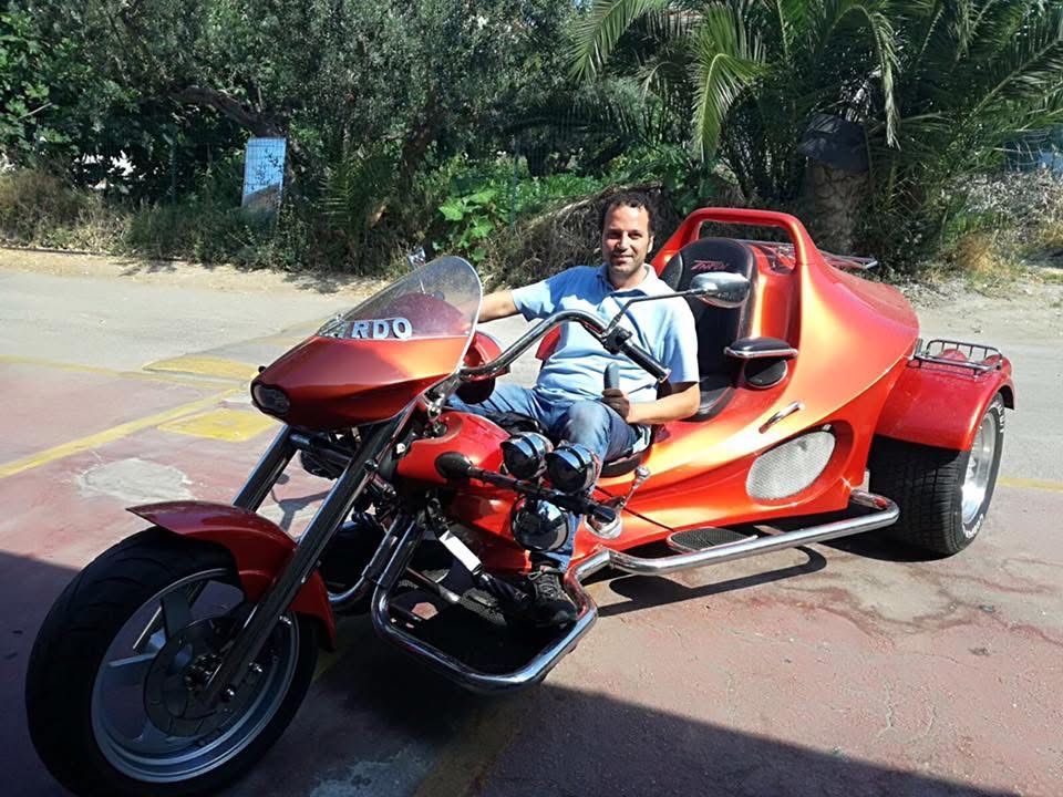 Uomo seduto su una motocicletta triciclo rossa, sorridente, all'aperto.