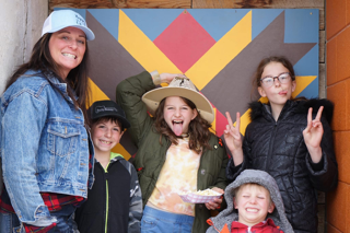 Group of five people smiling and making faces in front of a colorful geometric background.