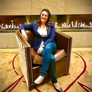 Woman in blazer and jeans sits in a brown armchair near a fireplace.