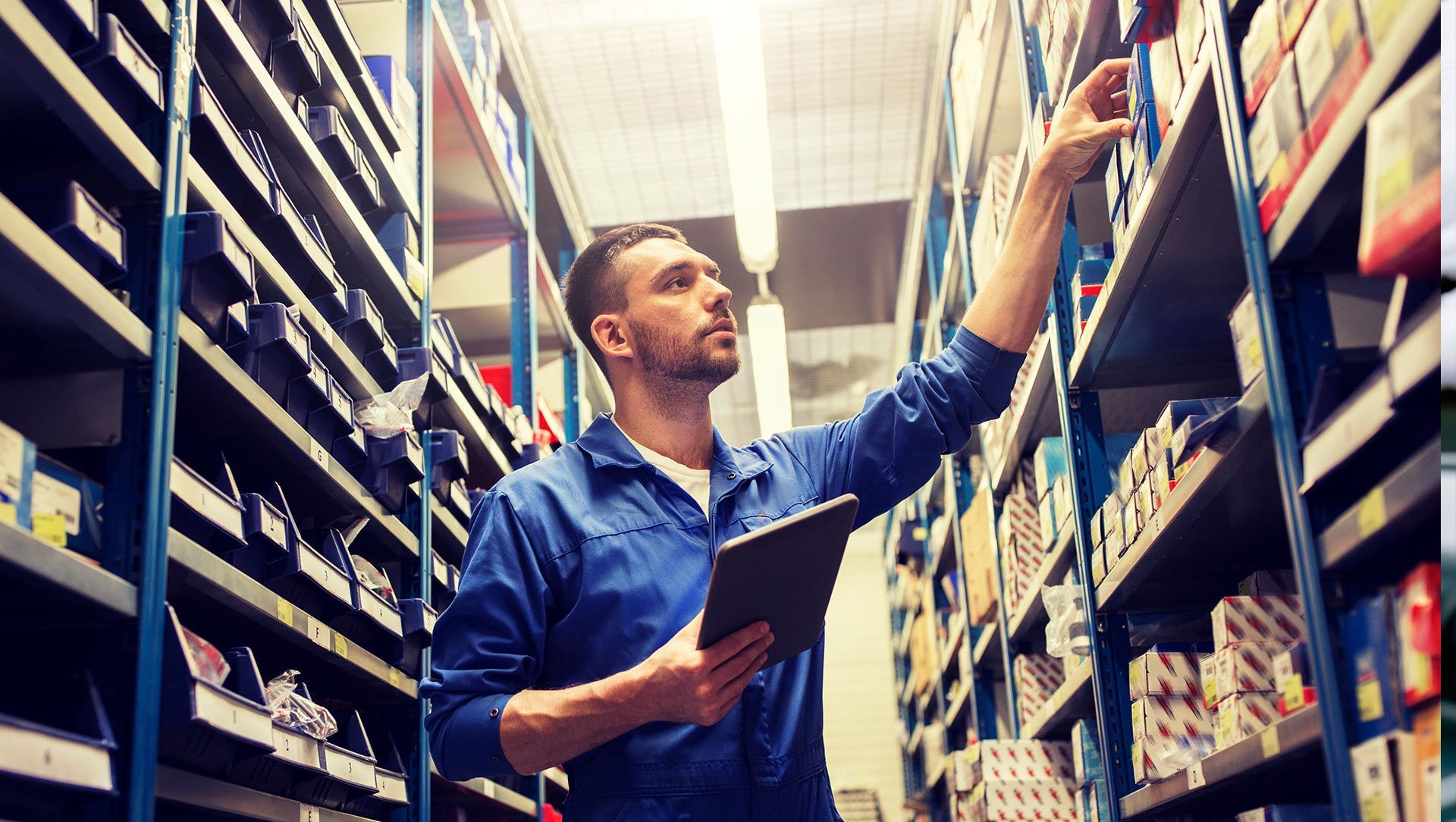 Man in blue jumpsuit reaching for a box on a shelf in a parts warehouse, holding a tablet.