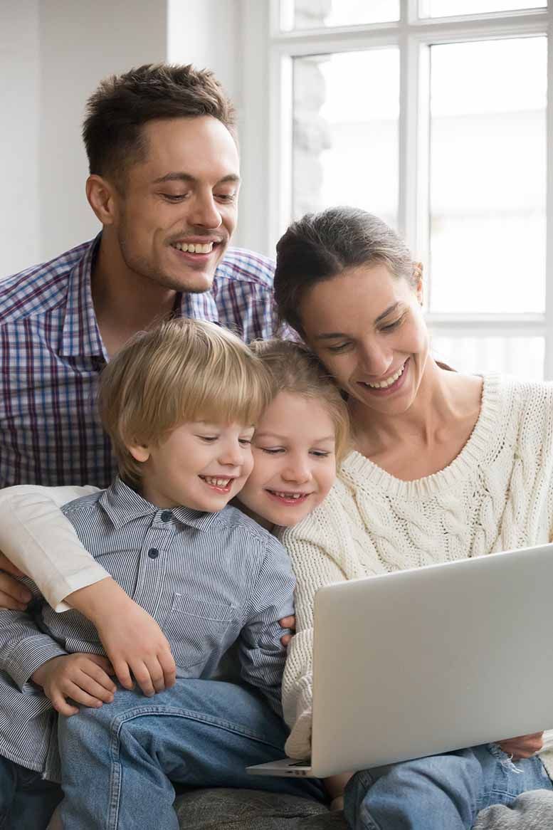 Family of four smiles while looking at a laptop computer. Window behind them.