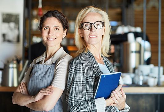 Two women, one in apron, one in blazer, smile with arms crossed in a cafe setting.