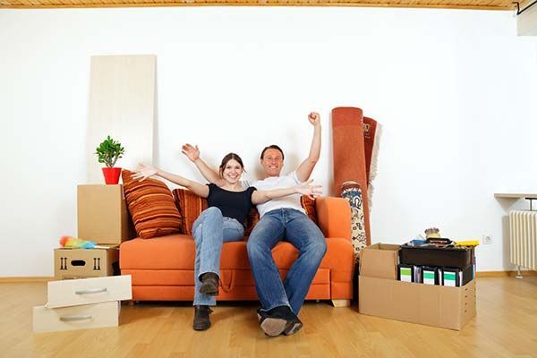 Couple cheers from orange couch in new empty apartment, surrounded by moving boxes and rolled-up rugs.