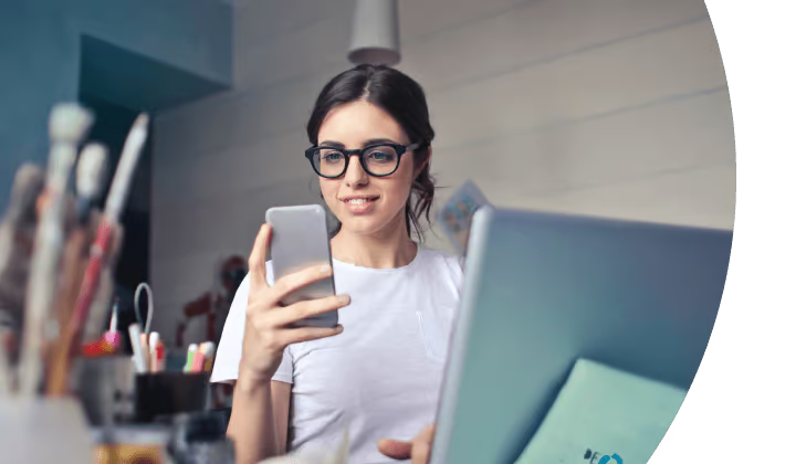Person with glasses using a smartphone at a desk with a laptop and brushes nearby