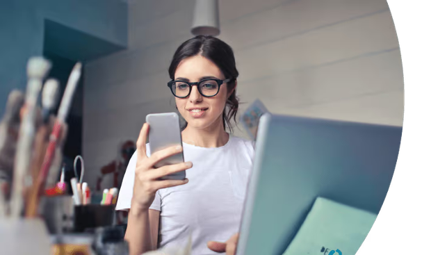 Person in glasses using a smartphone beside a laptop at a desk.