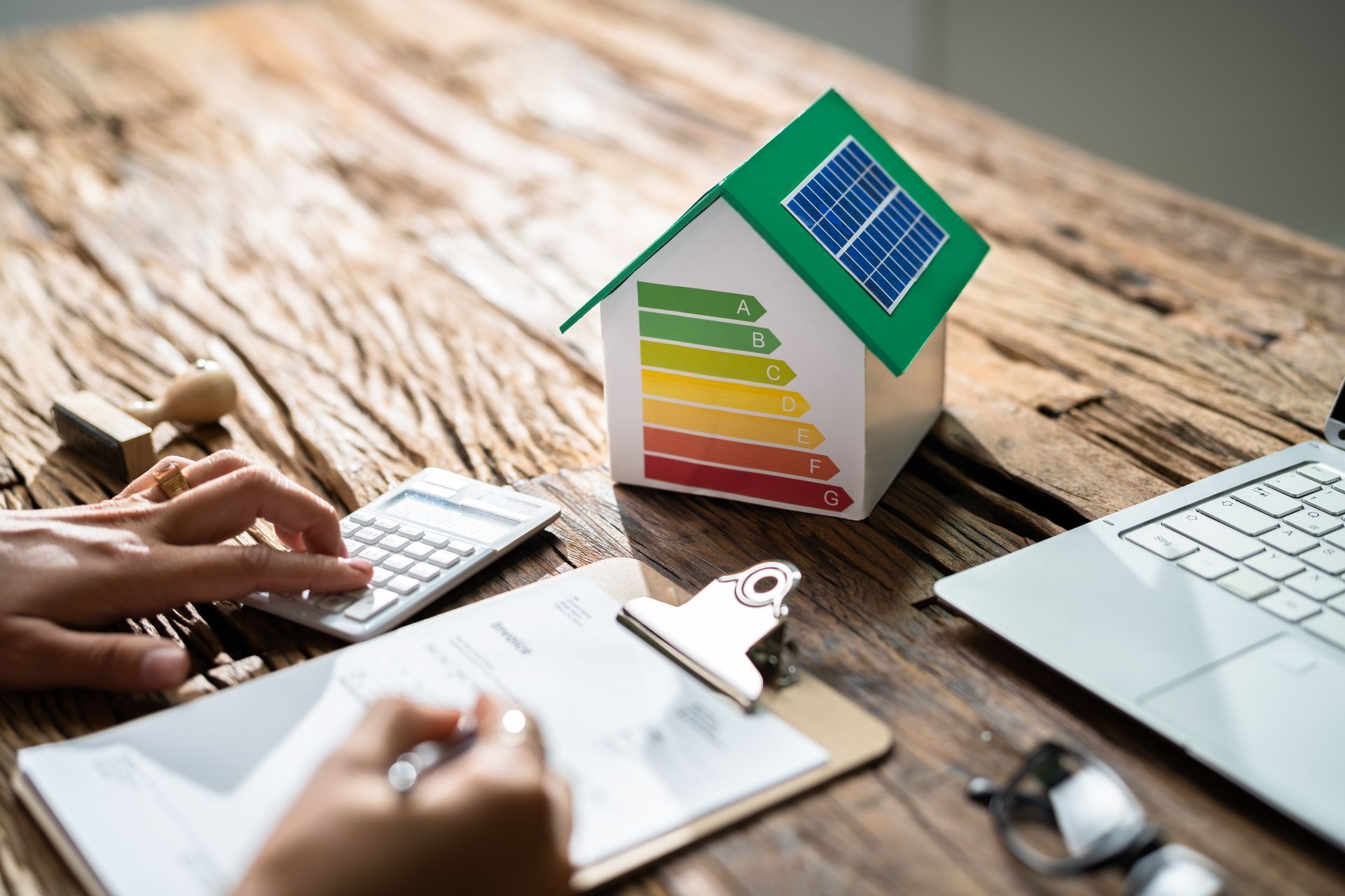 Hands using calculator and clipboard beside an energy-efficient house model and laptop on a desk