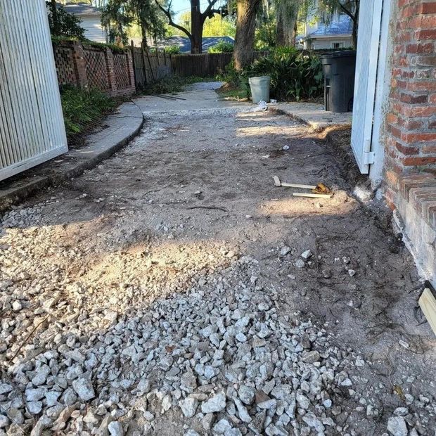 Gravel and dirt driveway under construction with a concrete border, leading to a fenced backyard.