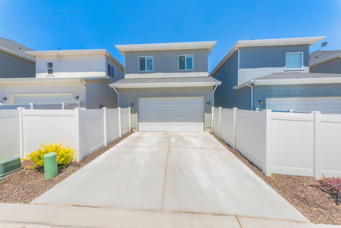 Two-story home with gray exterior, white garage door, and long concrete driveway between white fences on a sunny day.