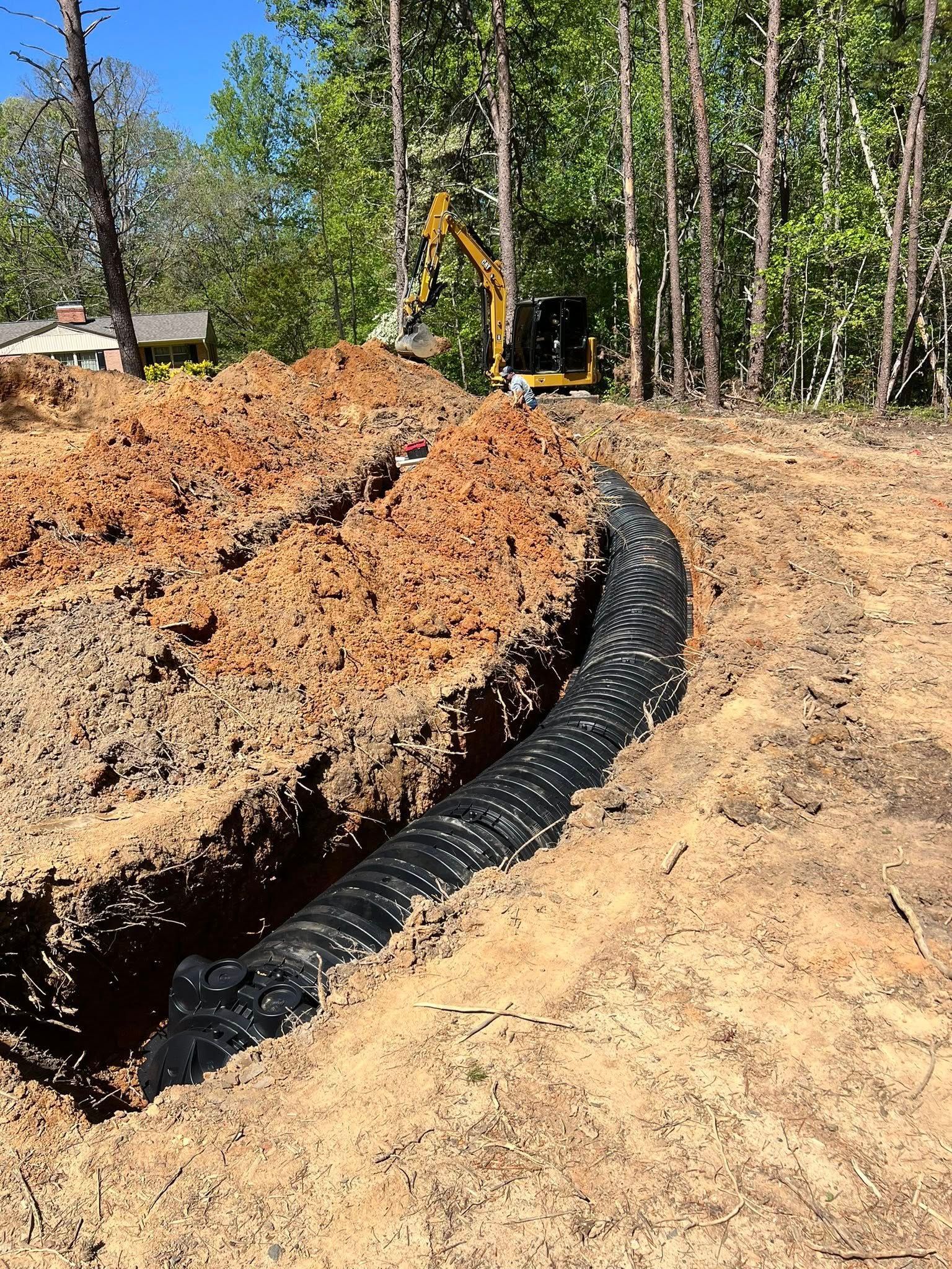A black corrugated drainage pipe sits in a dirt trench next to a mound of soil, with a yellow excavator in the background.