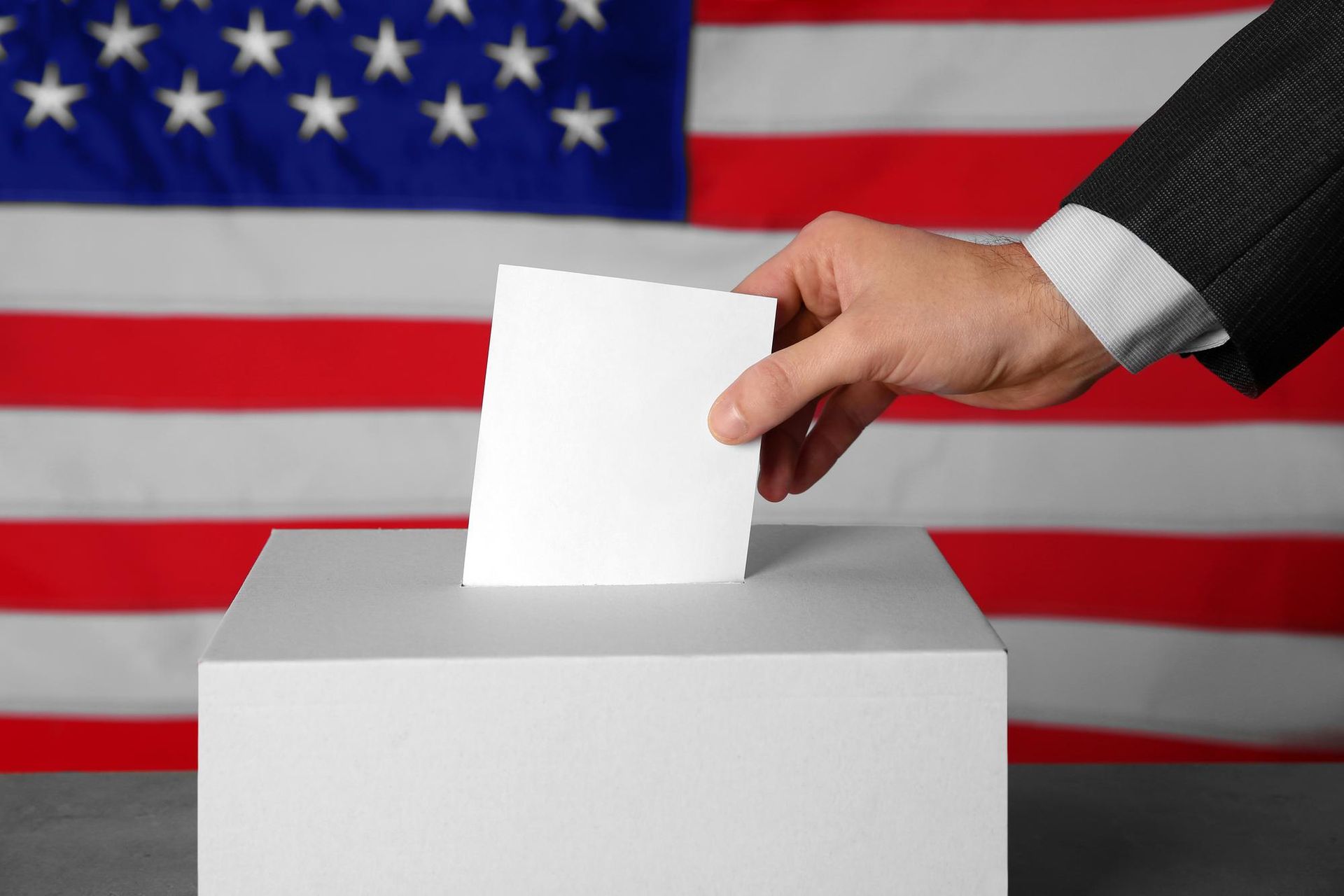 A person is putting a ballot into a ballot box in front of an american flag.