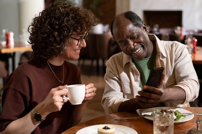 A man and a woman are sitting at a table drinking coffee and looking at a cell phone.