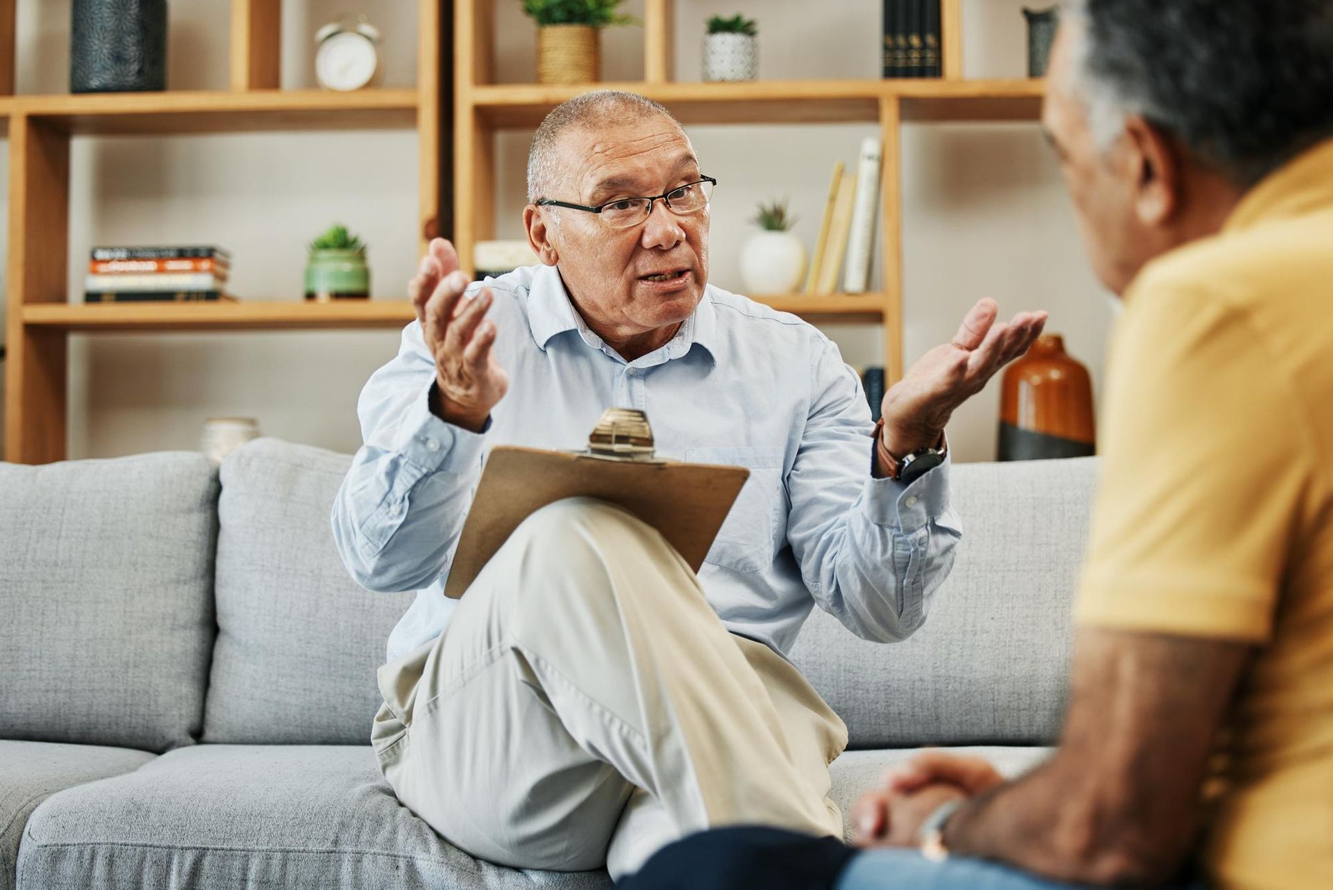 A man is sitting on a couch talking to another man while holding a clipboard.
