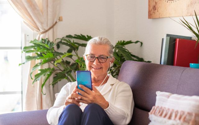 An elderly woman is sitting on a couch using a cell phone.