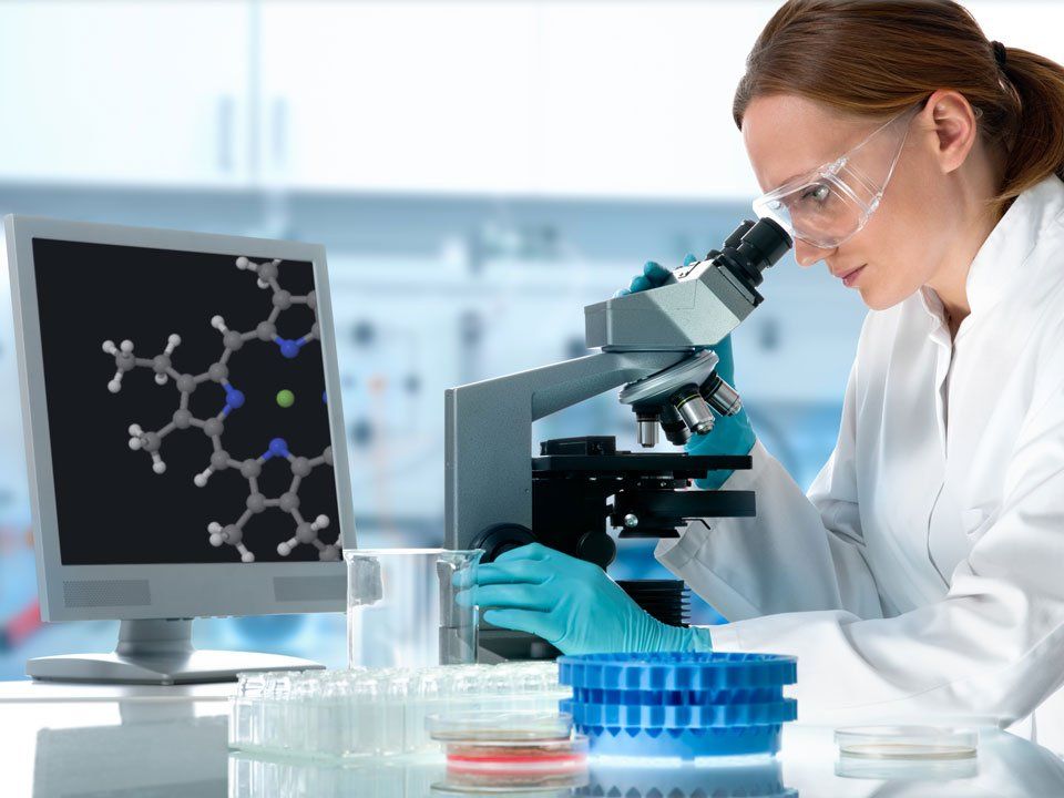 A woman is looking through a microscope in front of a computer screen