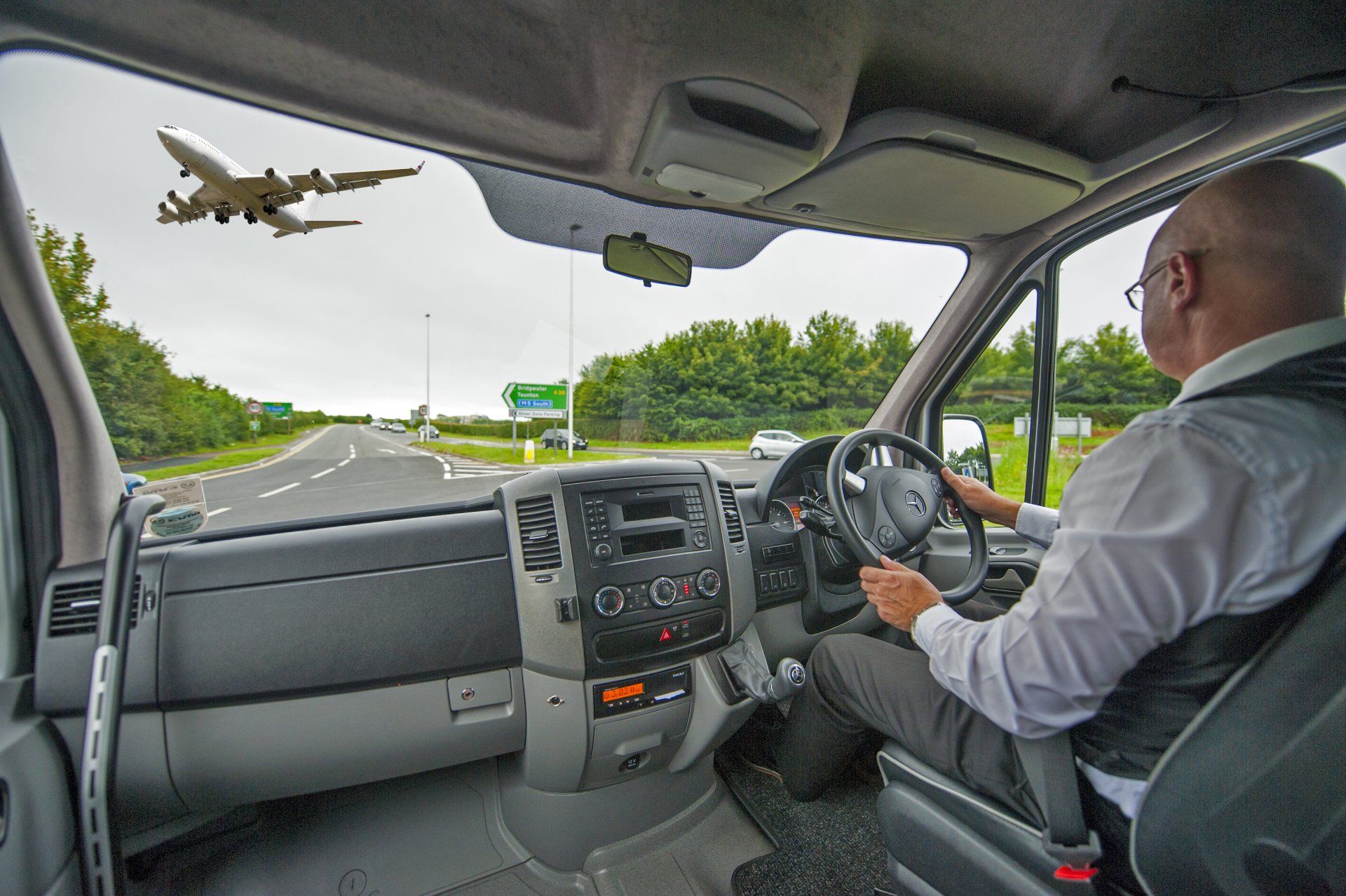 A man driving a van with a plane flying in the background