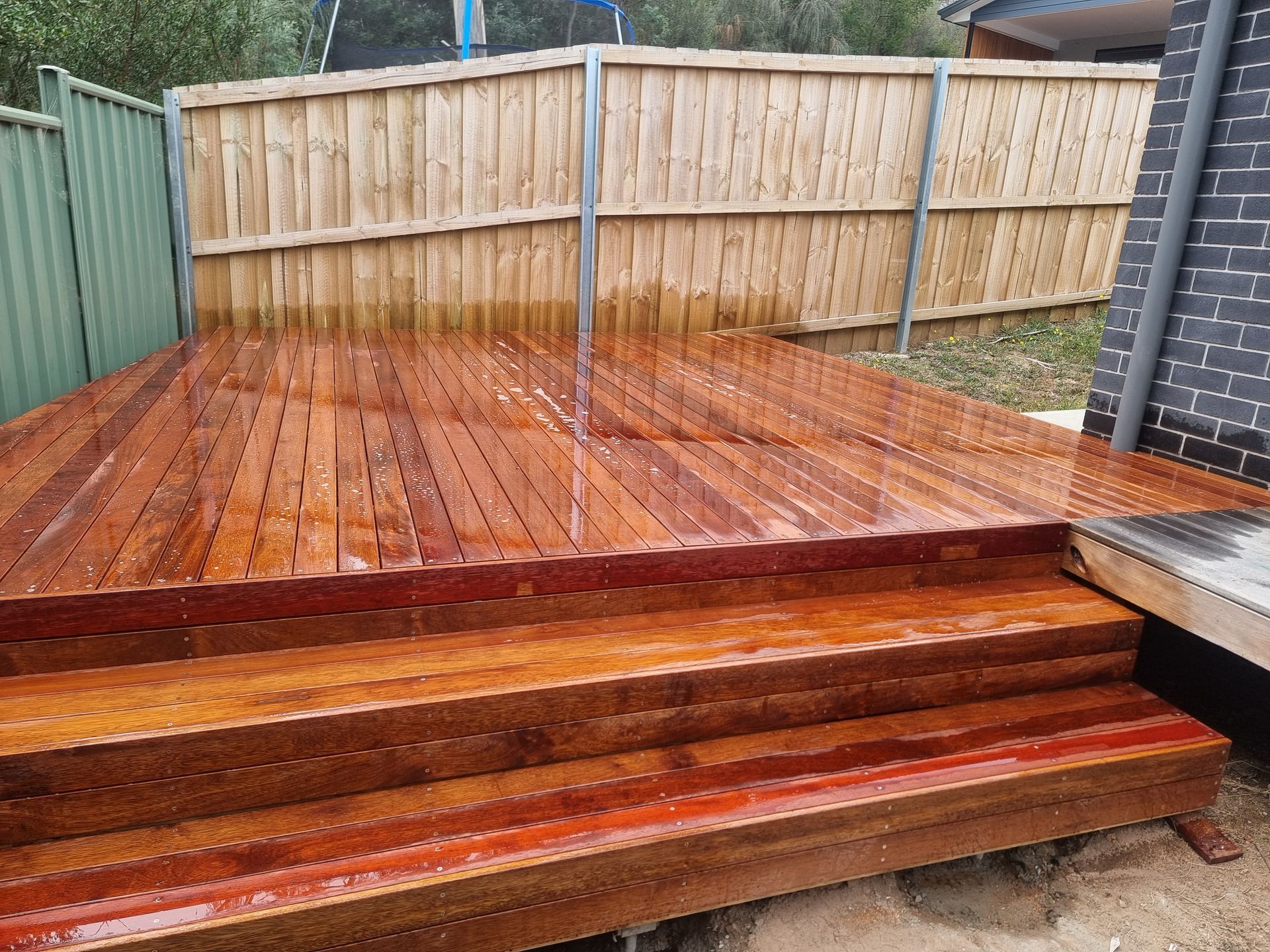 A freshly stained, wet wooden deck with two wide steps, situated outdoors against a wooden fence and a dark brick wall.