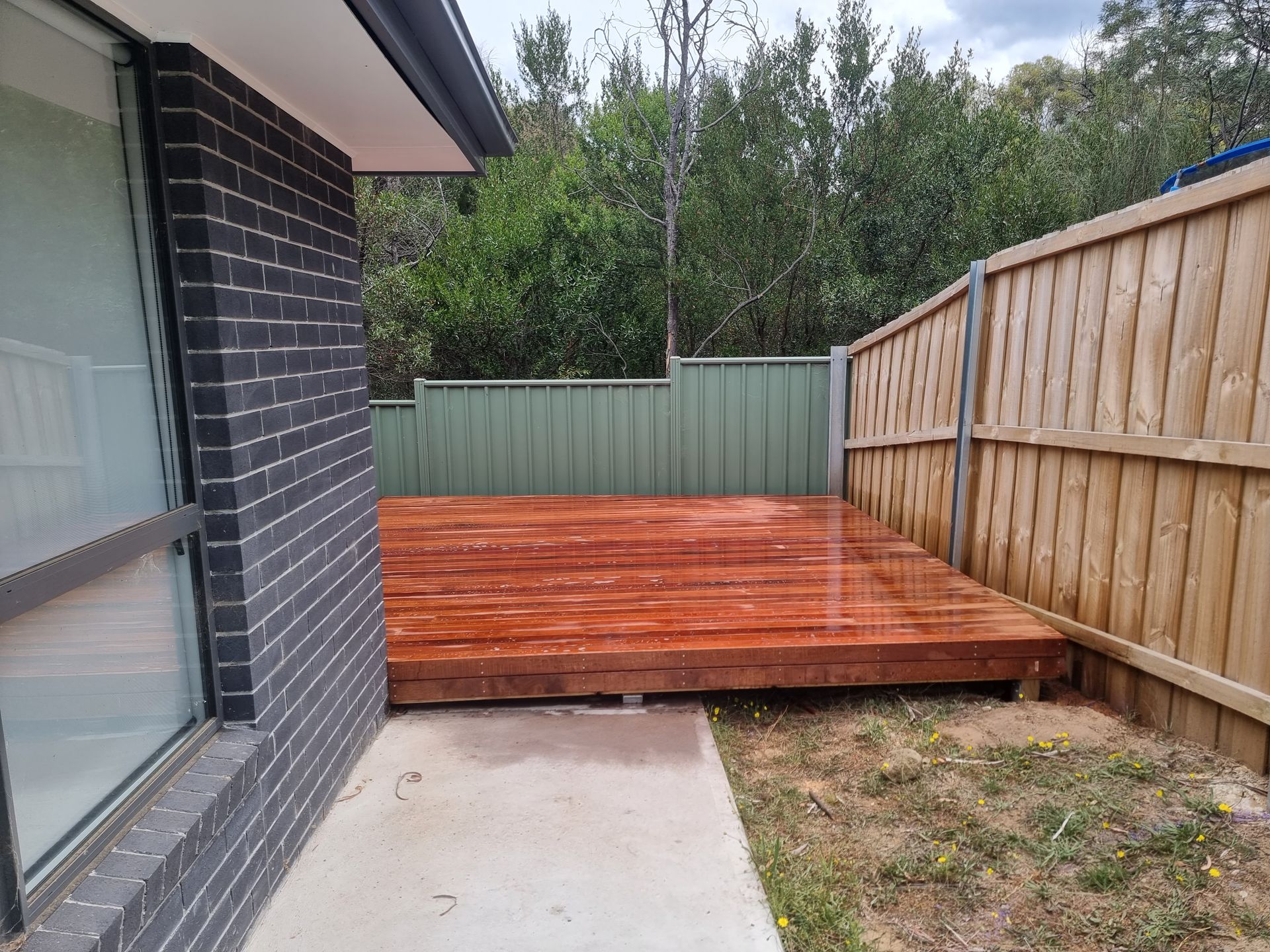 A newly stained rectangular wooden deck in a backyard, adjacent to a gray brick wall and a wooden fence.