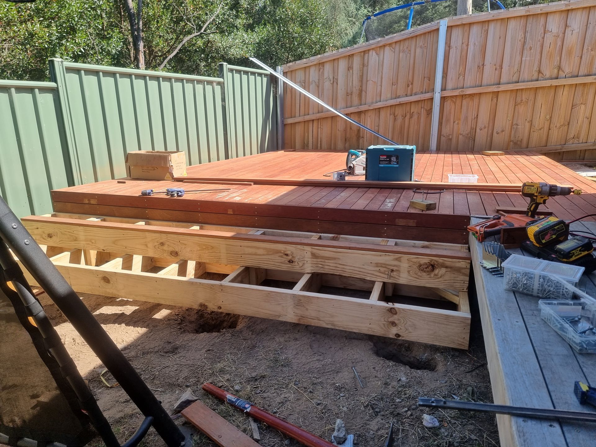A partially built wooden deck in a backyard with construction tools, framing joists, and installed planks.