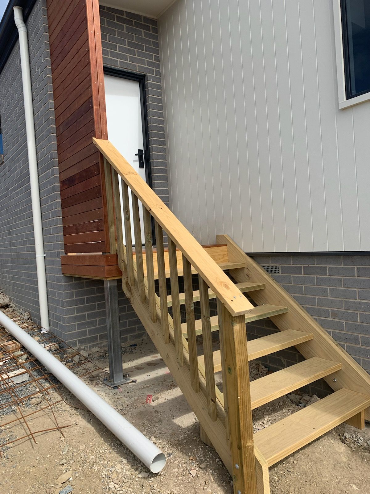 Wooden stairs with a handrail leading to a white door on the side of a house with grey brick and white siding.