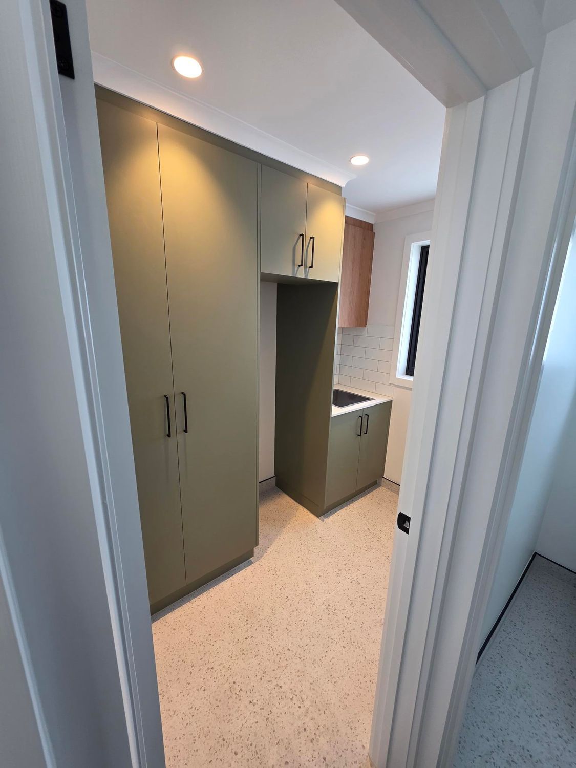 A laundry room with sage green cabinetry, a built-in sink, a small window, and speckled flooring seen through a doorway.