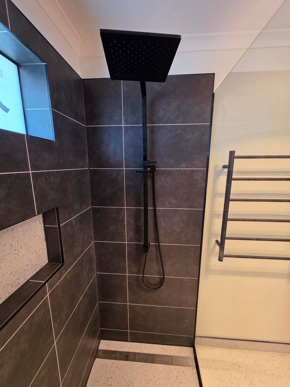 A black square rainfall shower head and handheld sprayer mounted on dark tiled walls next to a metal towel rack.