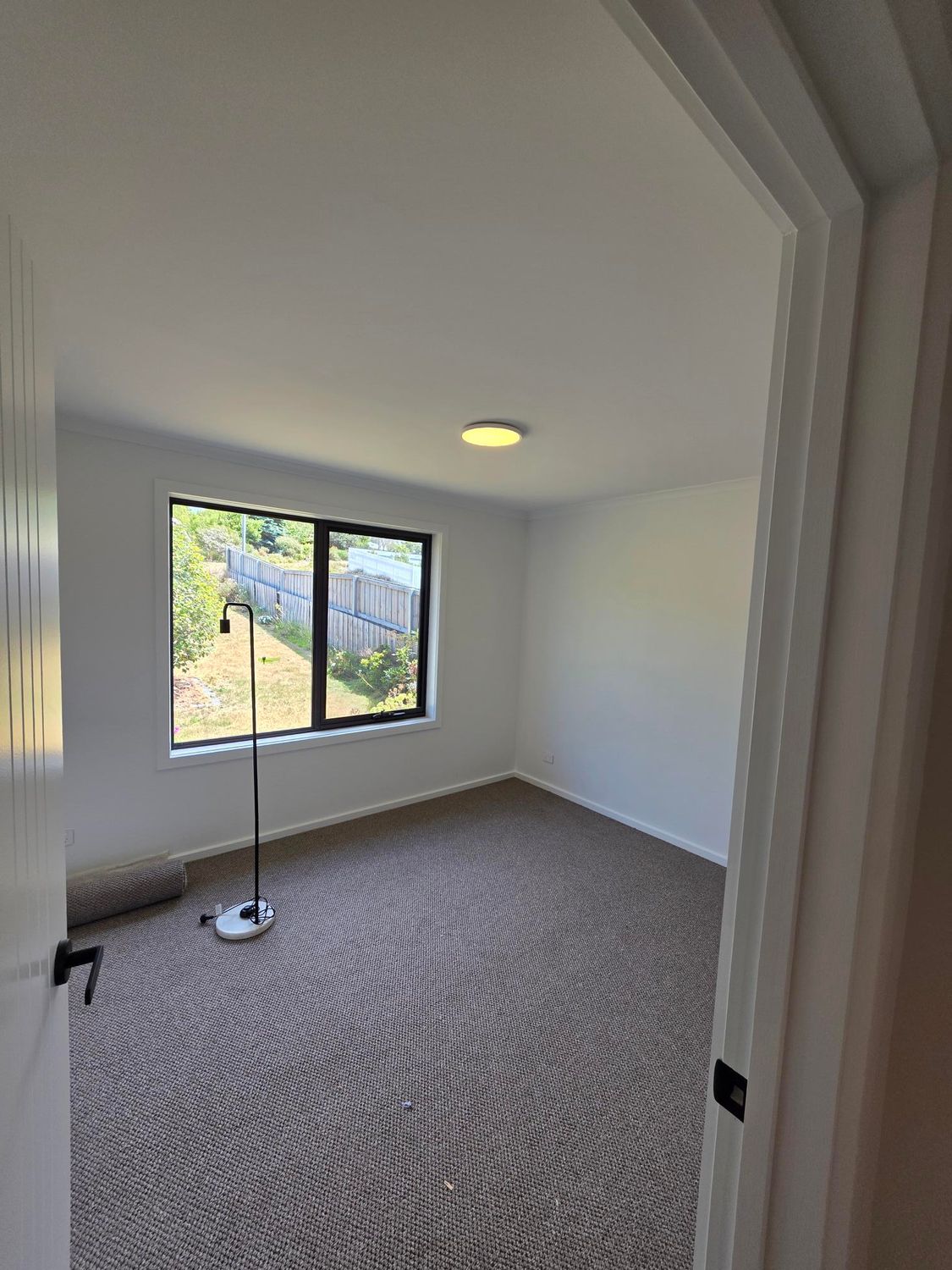 An empty room with light grey walls, patterned wall-to-wall carpet, a ceiling light, and a window with a yard view.