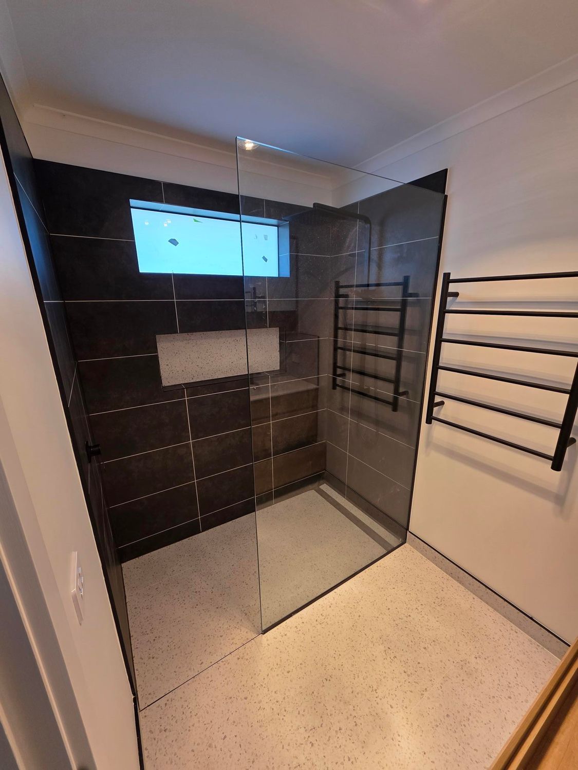 A modern walk-in shower with dark tiled walls, a light speckled floor, a glass partition, and black towel racks.