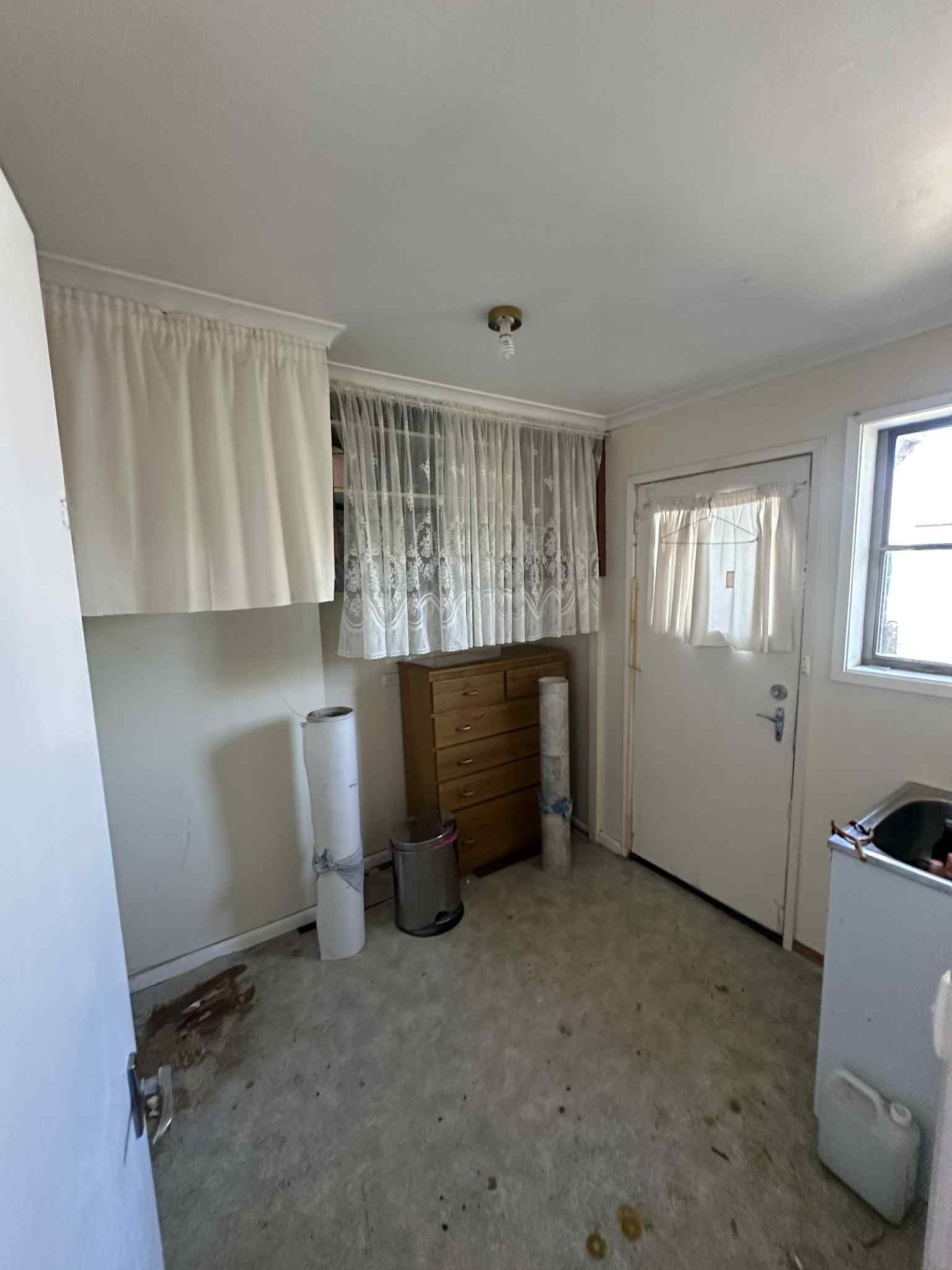 A sparsely furnished, utilitarian room with light walls, patterned curtains, a wooden dresser, and a door with a window.