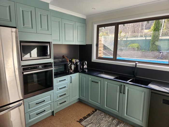 A kitchen with green cabinets , black counter tops , stainless steel appliances and a large window.