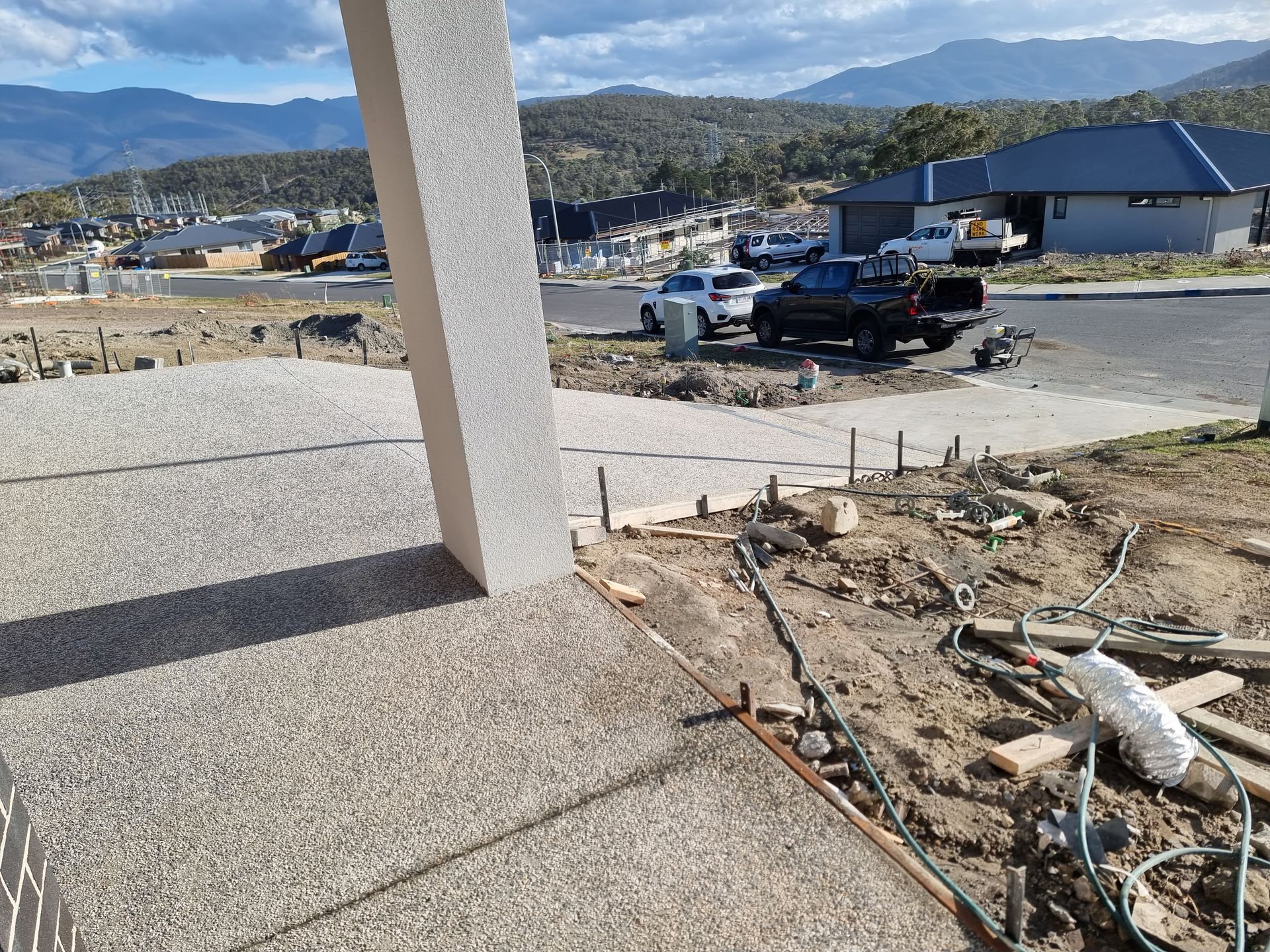 A sunlit construction site featuring a textured porch pillar and unfinished landscape area overlooking a suburban street.
