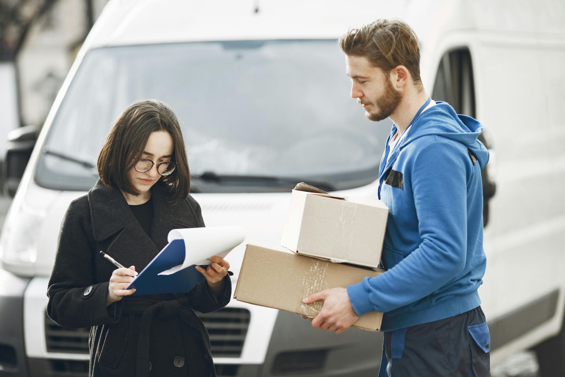 Two people reviewing papers beside a van, with one holding cardboard boxes
